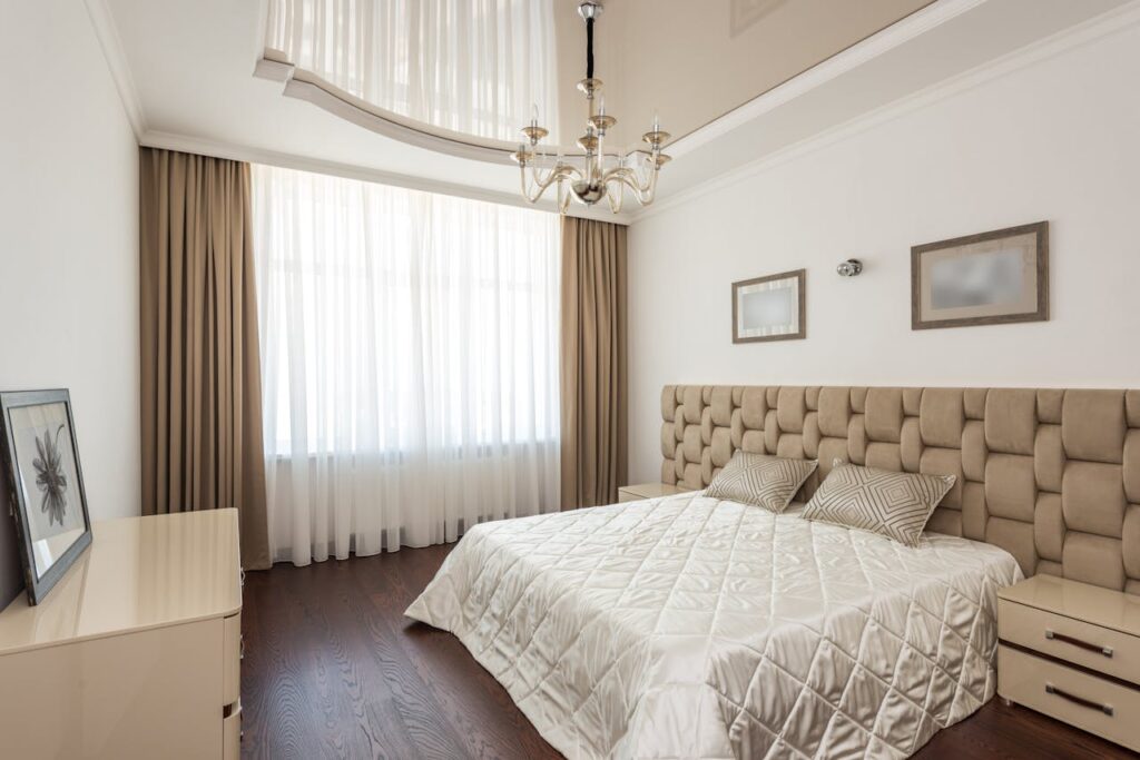 A modern bedroom featuring a luxurious bed, chandelier, and elegant decor with neutral tones.